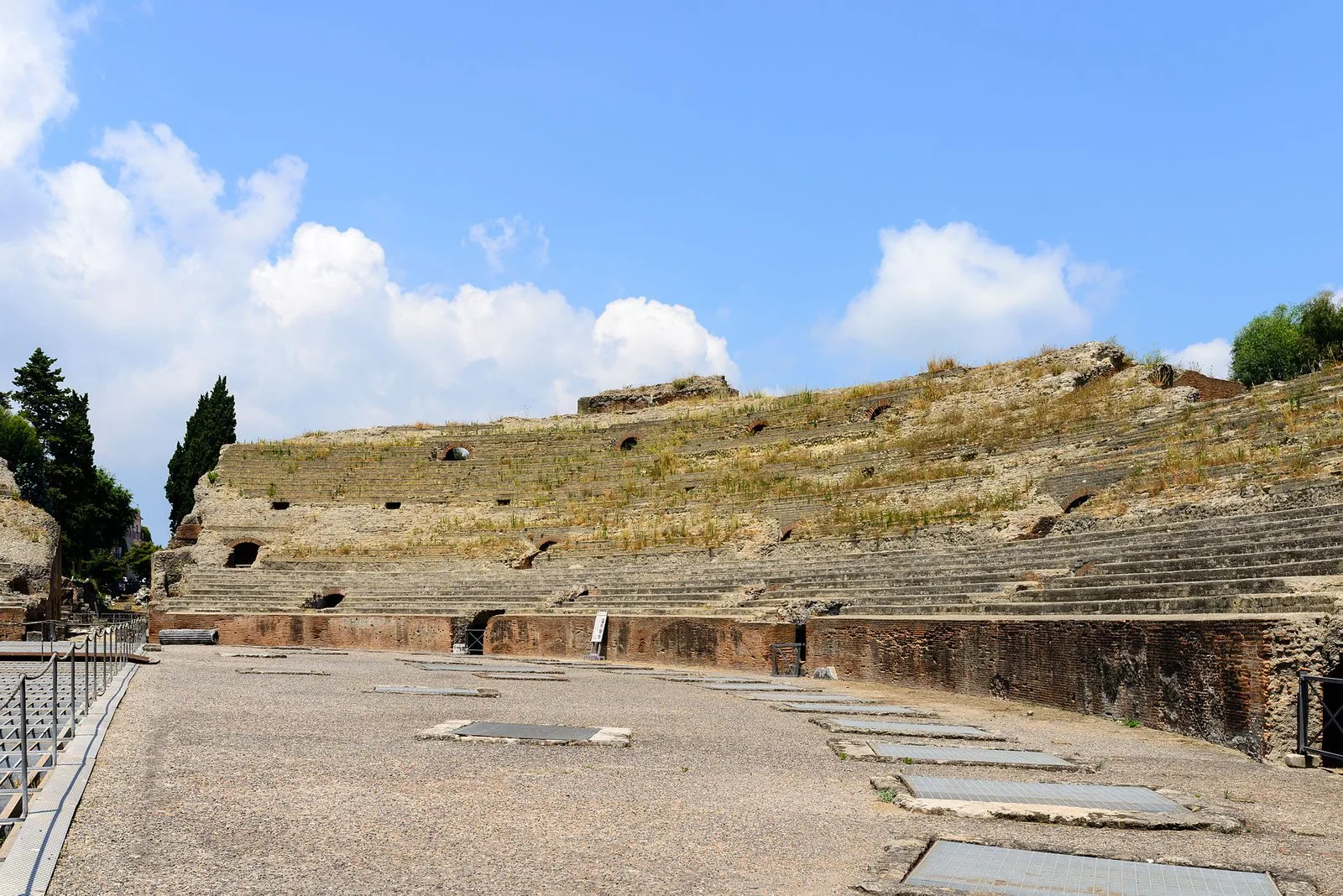 L'Anfiteatro Flavio di Pozzuoli: l'arena in terra battuta con le grate dei sotterranei, il muro di mattoni del podio e le gradinate in pietra coperte d'erba, con cipressi a sinistra e cielo azzurro con nuvole bianche