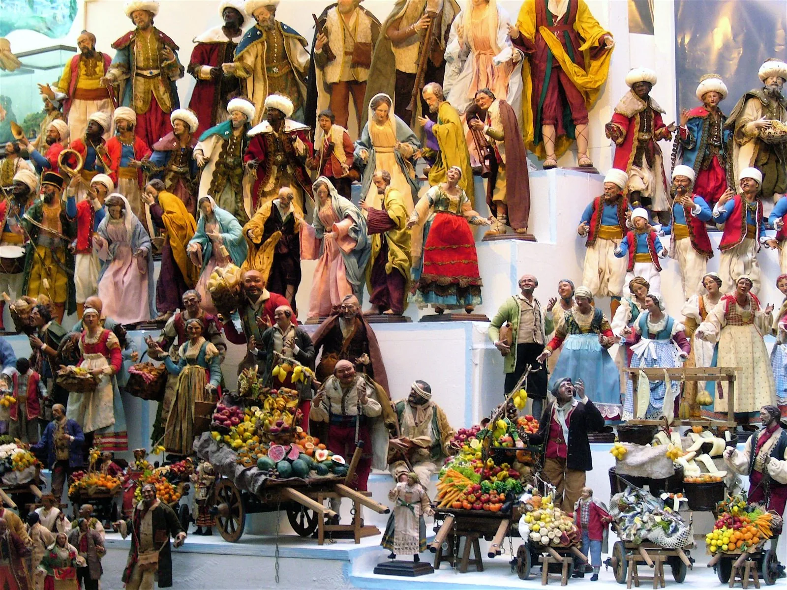 Banco di una bottega presepiale napoletana fitto di statuine: Re Magi con turbanti, la Madonna, pastori, musicisti e venditori con carretti carichi di frutta e verdura in miniatura