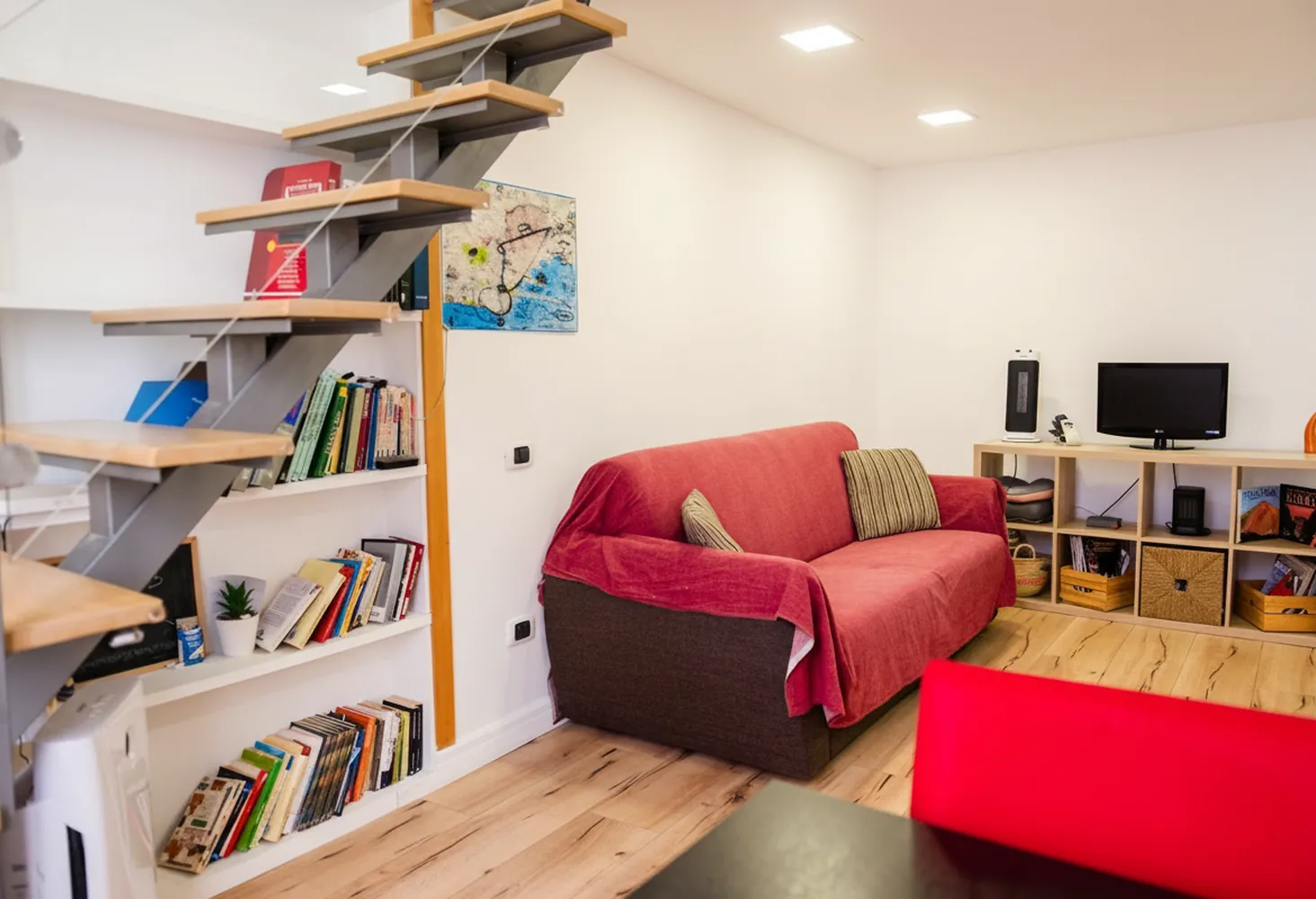 Il soggiorno con il divano letto rosso, la libreria sotto la scala e la TV sul mobile a giorno